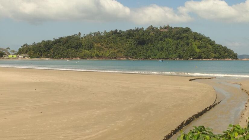 No canto direito, a calmaria domina: areia tranquila, casas ao redor e sombra sob as amendoeiras