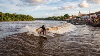 Uma fotografia de ação dinâmica, capturada ao nível dos olhos e ligeiramente em ângulo baixo, documenta o encontro único entre o homem e a natureza no Rio Mearim, próximo à cidade de Arari, Maranhão. O foco central é um surfista brasileiro experiente, com a pele dourada pelo sol e cabelo encaracolado, vestindo um short de surfe colorido. Ele está em pé sobre uma prancha, manobrando com habilidade a crista espumosa da pororoca, que se forma como uma onda marrom e turbulenta de sedimentos e água do mar. A onda avança poderosamente pelo rio, criando padrões circulares e redemoinhos complexos e escuros na água lamacenta ao redor do surfista. No fundo, à direita, a orla de Arari se estende ao longo da margem. Ela apresenta fileiras de casas coloniais coloridas com telhados de cerâmica vermelha e a torre de uma igreja branca, banhadas pela luz quente do fim da tarde. Dezenas de pessoas, locais e espectadores, estão reunidas nas escadas de pedra e nas plataformas de observação da orla, assistindo com entusiasmo à passagem do surfista e da onda. Pequenos barcos de pesca de madeira tradicionais estão ancorados perto da costa. No lado esquerdo do rio, a selva tropical densa e exuberante e a vegetação ribeirinha se estendem até a linha do horizonte sob um céu vasto e dramático com nuvens esparsas, iluminado pela luz dourada e suave do sol. A foto tem uma sensação de documentário autêntica e granulada, com cores vivas e foco nítido no surfista e na onda, capturando a energia crua e o momento singular