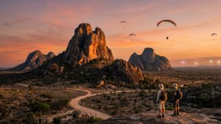 Uma fotografia de paisagem de grande angular, dramática e imersiva, captura a beleza sobrenatural de Quixadá, Ceará, durante o pôr do sol. A cena é dominada pelos maciços e únicos inselbergs, ou monólitos, que se elevam abruptamente da planície árida da Caatinga. A icônica formação "Pedra da Galinha Choca" ganha destaque no centro-esquerdo, iluminada pela luz quente e dourada do sol poente, que projeta sombras longas e profundas pelas rochas. As formações rochosas são compostas por quartzo-monzonito escuro e desgastado. No primeiro plano, uma trilha de terra sinuosa leva em direção à base dos monólitos, com vegetação resistente do deserto e cactos espalhados pelo terreno acidentado. Dois turistas de aventura, equipados com mochilas e vestindo roupas práticas para atividades ao ar livre (botas de caminhada, chapéus de abas largas), estão posicionados em um mirante no primeiro plano, pequenos contra a escala imensa das formações rochosas, olhando para o céu. Acima deles, alguns parapentes coloridos sobrevoam graciosamente o céu tingido de tons ricos de laranja, rosa e roxo, enfatizando a conexão da região com o voo e a energia. Ao longe, algumas luzes sutis, levemente anômalas e fracas, aparecem na borda do horizonte, adicionando um toque do folclore místico e ufológico da área sem ser explícito. O ar é límpido, a atmosfera transmite uma sensação de reverência e aventura silenciosa. A luz é difusa, criando uma experiência visual majestosa e poderosa. A imagem tem granulado de filme e uma paleta de cores rica, em orientação paisagem.