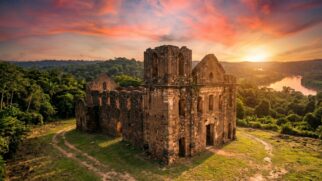 Uma fotografia panorâmica de grande angular e cinematicamente dramática captura as imponentes e solitárias ruínas da Igreja de Nossa Senhora do Rosário dos Pretos em Vila Bela da Santíssima Trindade, Mato Grosso, durante um pôr do sol espetacular. A cena é dominada pela estrutura remanescente da igreja inacabada do século XVIII, cujas massivas paredes de pedra e adobe desgastadas se erguem centralmente contra um céu vibrante, preenchido por nuvens texturizadas em tons intensos de laranja, rosa, roxo e dourado. A luz do sol poente, vinda da direita, banha a fachada de pedra escura e texturizada com um brilho dourado e quente, acentuando o desgaste e os detalhes arquitetônicos dos arcos e aberturas, que revelam a ausência de telhado e o interior oco. A igreja está situada em uma clareira de grama verde-esmeralda e caminhos de terra batida que serpenteiam em direção à estrutura. Ao redor da clareira, uma selva tropical densa e exuberante envolve as ruínas, criando uma moldura verde natural que se estende até colinas onduladas na distância. Ao fundo, à direita, o Rio Guaporé sinuoso reflete as cores do pôr do sol e flui através da floresta até o horizonte. A atmosfera é de solitude, mistério e grandeza histórica, com o passado colonial abandonado à majestade da natureza selvagem. A foto possui granulado de filme natural, cores ricas e foco nítido na igreja e na vegetação ao redor.