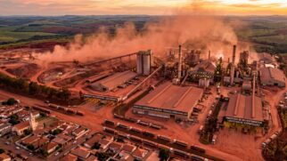 Uma foto aérea panorâmica captura um vasto complexo industrial de mineração e fabricação de cerâmica no Brasil, operando dia e noite, ao pôr do sol. Toda a cena é banhada por um tom avermelhado generalizado, desde os estoques de matéria-prima até as estradas de terra, enfatizando a escala da operação. A instalação inclui fábricas extensas, inúmeras chaminés emitindo fumaça e sistemas complexos de correias transportadoras. Abaixo, uma longa fileira de semi-caminhões aguarda para carregar. Uma pequena área residencial com uma igreja está localizada logo ao lado do complexo industrial. Um grande cartaz identifica o local como a 'Mineração Santa Gertrudes', o Maior Polo Cerâmico das Américas, destacando seu papel como uma indústria para revestimentos e logística de exportação.