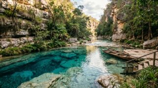 Uma fotografia de paisagem horizontal, em ângulo médio e ao nível dos olhos, captura uma cena serena em um cânion cárstico isolado no cerrado brasileiro. O foco principal é uma piscina natural de água incrivelmente límpida e cristalina, que exibe tons vibrantes de azul-turquesa e safira. A transparência da água permite a visualização detalhada do leito do rio, composto por formações rochosas calcárias e pequenas pedras. O poço é emoldurado por altas e imponentes falésias de calcário, texturizadas e desgastadas, que se elevam em ambos os lados, cobertas por uma vegetação densa e variada, incluindo samambaias, arbustos e árvores nativas do cerrado. À direita, um simples deque de madeira com escadas e corrimão leva à água, onde duas canoas de madeira tradicionais estão atracadas. A luz solar suave da tarde filtra-se do topo do cânion, iluminando a água e as plantas, criando reflexos e uma atmosfera mágica e escondida.