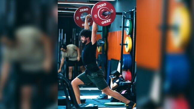 Homem realizando levantamento de peso olímpico em uma academia. Ele segura uma barra com anilhas vermelhas acima da cabeça, em posição de avanço (split jerk), vestindo camiseta preta e bermuda escura. Ao fundo, há equipamentos de treino e outras pessoas desfocadas.