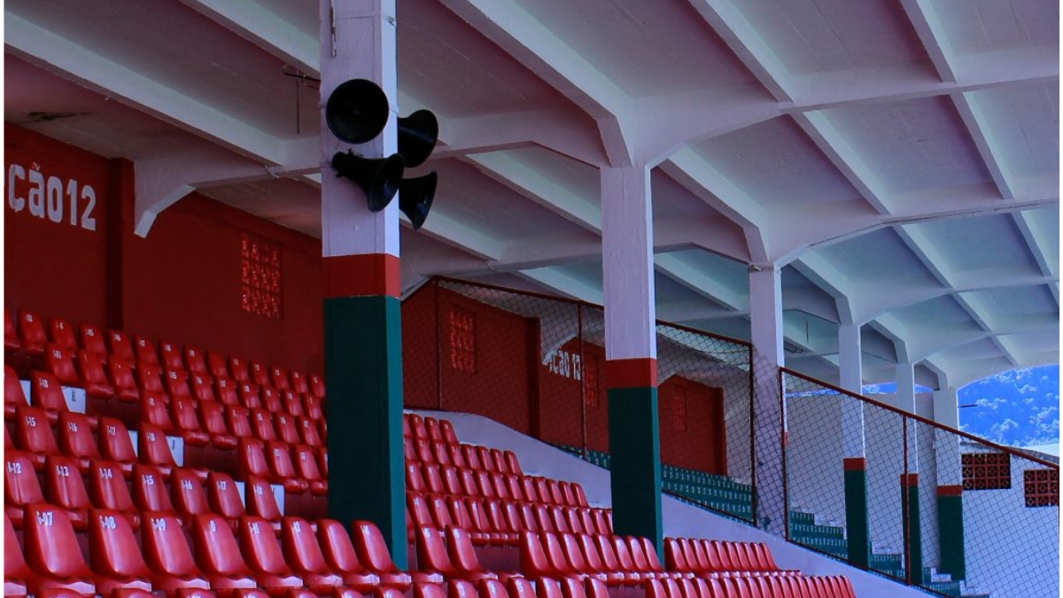 Arquibancada com cadeiras vermelhas e estrutura coberta em estádio, com pilares nas cores verde, vermelho e branco.