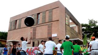 Grupo de pessoas tocando e se apresentando em frente a prédio de tijolos com grafite ao fundo.