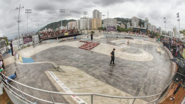 Pista de skate estilo street com skatistas andando, arquibancadas cheias e prédios ao fundo sob céu nublado.