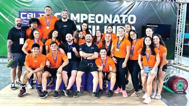 Grupo de atletas e equipe técnica posando para foto em um pódio de competição. A maioria veste camisetas laranjas, alguns com medalhas no peito, enquanto outros usam camisetas pretas. Ao fundo, um painel verde com o logotipo da CBLP e a escrita "Campeonato Brasileiro". Anilhas de peso são visíveis no canto inferior direito.