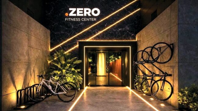 Fachada da academia Zero Fitness Center com iluminação em fitas de LED amarelas no chão e nas paredes de concreto aparente. À esquerda, bicicletas estão estacionadas em um suporte, e à direita, outras bicicletas estão penduradas na parede. O logotipo da academia está centralizado no topo sobre uma parede de mármore escuro.