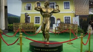 Estátua de bronze do fisiculturista Arnold Schwarzenegger em uma pose de exibição de músculos (double biceps). A escultura está sobre uma base circular com seu nome, cercada por cordões de isolamento vermelhos. Ao fundo, um painel fotográfico retrata uma casa amarela com janelas brancas, bancos de madeira e gramado sintético verde no chão.