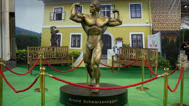 Estátua de bronze do fisiculturista Arnold Schwarzenegger em uma pose de exibição de músculos (double biceps). A escultura está sobre uma base circular com seu nome, cercada por cordões de isolamento vermelhos. Ao fundo, um painel fotográfico retrata uma casa amarela com janelas brancas, bancos de madeira e gramado sintético verde no chão.
