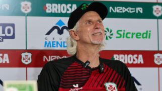 Homem de cabelos brancos e boné preto sorrindo durante uma entrevista coletiva. Ele veste uma camisa polo listrada em preto e vermelho com o escudo da Portuguesa Santista e utiliza microfones de lapela. Ao fundo, um painel de patrocinadores exibe marcas como Briosa, Sicredi e Grupo Mendes.