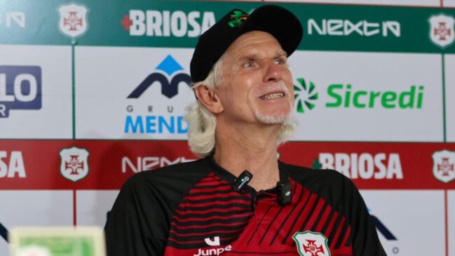 Homem de cabelos brancos e boné preto sorrindo durante uma entrevista coletiva. Ele veste uma camisa polo listrada em preto e vermelho com o escudo da Portuguesa Santista e utiliza microfones de lapela. Ao fundo, um painel de patrocinadores exibe marcas como Briosa, Sicredi e Grupo Mendes.