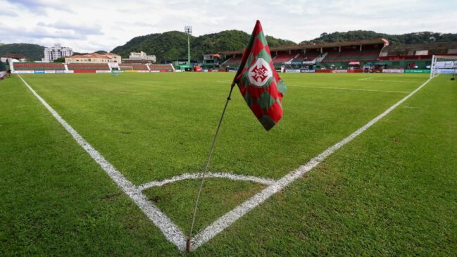 Bandeirinha de escanteio com escudo da Portuguesa Santista em campo de futebol, com arquibancadas ao fundo.