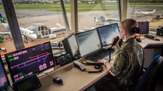A imagem captura o interior de uma torre de controle de tráfego aéreo a partir de um ângulo levemente elevado, destacando uma profissional em plena atividade. Em primeiro plano, uma controladora aparece de perfil e de costas para a câmera, vestindo uma farda militar camuflada e com o cabelo rigorosamente preso em um coque. Ela segura um microfone de rádio próximo ao rosto com a mão direita, indicando um momento de comunicação ativa, enquanto sua mão esquerda se apoia sobre uma extensa bancada de trabalho repleta de tecnologia. Sobre o console, destacam-se três monitores principais que exibem informações críticas: o da esquerda apresenta um mapa de radar colorido com o traçado das pistas, enquanto os centrais mostram dados técnicos e tabelas de voo. Além das telas, nota-se a presença de um teclado preto, mouse e diversos módulos de comunicação com cabos espiralados que reforçam a complexidade da função. Ao fundo, através das amplas janelas de vidro que circundam a torre, é possível ver o pátio de um aeroporto sob a luz clara do dia. Uma aeronave da companhia Azul está estacionada e conectada a uma ponte de embarque, cercada pela infraestrutura asfáltica e áreas verdes do sítio aeroportuário. A composição geral transmite uma atmosfera de vigilância, precisão técnica e segurança operacional.