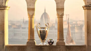 A icônica taça da UEFA Champions League e a bola oficial da Adidas posicionadas em uma mureta de arcos de pedra. Ao fundo, destaca-se a arquitetura clássica de Budapeste, com o Parlamento Húngaro sob uma luz suave de entardecer.