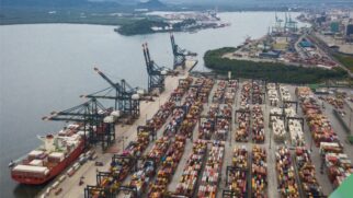 Esta fotografia aérea apresenta uma visão panorâmica e detalhada de um terminal de contêineres em plena operação no Porto de Santos. A imagem é composta por elementos que reforçam a magnitude e a organização do setor logístico na região. No primeiro plano, à esquerda, observa-se um grande navio cargueiro atracado, carregado com diversas camadas de contêineres coloridos. Ao longo do cais, diversas pontes rolantes (portêineres) de grande porte, pintadas de azul e preto, estão posicionadas para a movimentação das cargas entre o navio e o pátio. O pátio do terminal ocupa a maior parte da composição, exibindo milhares de contêineres organizados em pilhas precisas, criando um mosaico de cores como vermelho, azul, branco e amarelo. Entre as fileiras, é possível notar a infraestrutura de apoio, como guindastes de pátio e carretas que realizam o transporte interno das mercadorias. Ao fundo, a paisagem é composta pelo canal do porto, áreas de vegetação nativa (manguezais) e a silhueta de outros terminais e edifícios da cidade sob um céu nublado. A luz natural difusa destaca os detalhes industriais sem criar sombras pesadas, conferindo à imagem um tom profissional e realista, ideal para ilustrar reportagens sobre economia, logística e o mercado de trabalho portuário.