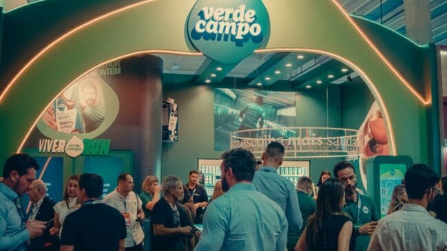 Estande da Verde Campo em evento fitness, com design moderno em tons de verde, iluminação em arco e visitantes interagindo com produtos e painéis informativos sobre nutrição e bem-estar.