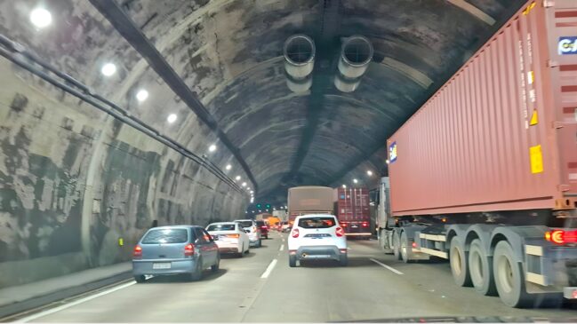 Uma fotografia de rua realista capturada do ponto de vista de um motorista mostra o tráfego parado dentro de um túnel de rodovia movimentado. O túnel é construído com paredes de concreto áspero e possui uma forma circular em arco, pontuada por luzes de teto redondas e brilhantes instaladas ao longo de toda a extensão do teto do túnel. Dois grandes ventiladores de metal estão pendurados no teto, perto do meio da imagem. À direita, um grande caminhão de contêiner marrom ocupa toda a faixa, com a parte traseira de seu contêiner marrom texturizado, pneus grandes e sistema de reboque dominando o primeiro plano à direita. Em seguida, uma fileira de carros de passageiros brancos e prateados está alinhada em tráfego intenso na faixa esquerda do túnel, incluindo um carro compacto prateado, um sedan branco e um SUV branco, com as luzes de freio vermelhas acesas. Um pequeno carro prateado é visto no primeiro plano à esquerda. O teto e as paredes do túnel são feitos de concreto texturizado e desgastado, com tubulações expostas, eletrodutos e fiação visíveis ao longo da parede direita. A iluminação de teto redonda e brilhante cria uma série de pontos de luz quentes e uniformemente espaçados ao longo do teto arqueado do túnel. O asfalto da rodovia é desgastado e possui marcas de pista brancas e contínuas que se estendem para a distância. O tráfego parado continua para o túnel profundo na distância, com mais luzes traseiras vermelhas brilhantes e veículos indistintos visíveis.