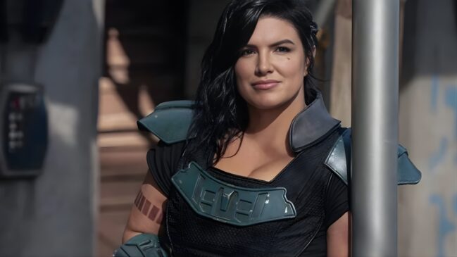 A imagem apresenta a atriz Gina Carano caracterizada como sua personagem Cara Dune, da série "The Mandalorian". Ela aparece em um plano médio, com uma expressão serena e um leve sorriso, olhando diretamente para a câmera. Seus cabelos são pretos, longos e levemente ondulados, caindo sobre os ombros. Ela veste o traje de combate característico da personagem, composto por uma armadura peitoral escura com detalhes em verde-azulado metálico e ombreiras robustas no mesmo tom. No braço direito, é possível observar uma tatuagem de barras horizontais, que faz parte do figurino da ex-soldada. O fundo da cena é desfocado, sugerindo um ambiente externo de um posto avançado ou cidade do universo de Star Wars, com tons neutros e estruturas metálicas, enquanto um poste vertical prateado aparece em primeiro plano à direita, emoldurando a composição