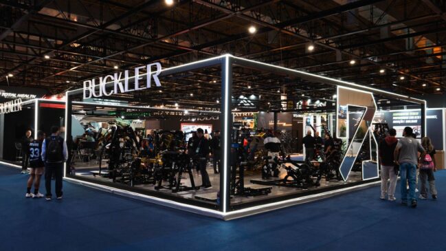 Estande da Buckler em feira, com estrutura iluminada em formato aberto, exposição de máquinas de musculação e visitantes circulando e testando os aparelhos.