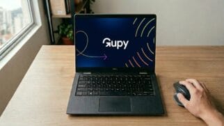 Esta fotografia em estilo corporativo e realista apresenta uma estação de trabalho moderna e organizada em um ambiente de home office banhado por luz natural. No centro da cena, um notebook Dell de cor preta está aberto sobre uma mesa de madeira clara, exibindo na tela a identidade visual vibrante da plataforma Gupy com seu logotipo branco e grafismos em degradê sobre um fundo azul-escuro. A composição inclui a mão de uma pessoa no canto inferior direito operando um mouse ergonômico de alta precisão, o que confere uma sensação de atividade e foco profissional ao cenário. Ao fundo, o desfoque suave revela uma estante com livros e uma janela com vista para uma paisagem urbana, criando uma atmosfera autêntica de trabalho remoto que equilibra tecnologia e conforto.