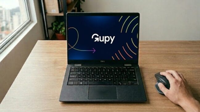 Esta fotografia em estilo corporativo e realista apresenta uma estação de trabalho moderna e organizada em um ambiente de home office banhado por luz natural. No centro da cena, um notebook Dell de cor preta está aberto sobre uma mesa de madeira clara, exibindo na tela a identidade visual vibrante da plataforma Gupy com seu logotipo branco e grafismos em degradê sobre um fundo azul-escuro. A composição inclui a mão de uma pessoa no canto inferior direito operando um mouse ergonômico de alta precisão, o que confere uma sensação de atividade e foco profissional ao cenário. Ao fundo, o desfoque suave revela uma estante com livros e uma janela com vista para uma paisagem urbana, criando uma atmosfera autêntica de trabalho remoto que equilibra tecnologia e conforto.