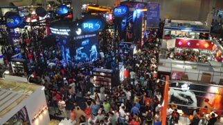 A imagem retrata o pavilhão do Arnold Sports lotado, com centenas de pessoas circulando entre grandes estandes de suplementação. Em destaque central, aparecem as estruturas da marca Probiótica, e no canto inferior, o estande da MuscleMeds, evidenciando o clima de grande evento de fisiculturismo.