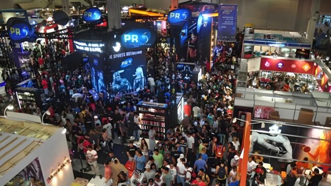A imagem retrata o pavilhão do Arnold Sports lotado, com centenas de pessoas circulando entre grandes estandes de suplementação. Em destaque central, aparecem as estruturas da marca Probiótica, e no canto inferior, o estande da MuscleMeds, evidenciando o clima de grande evento de fisiculturismo.