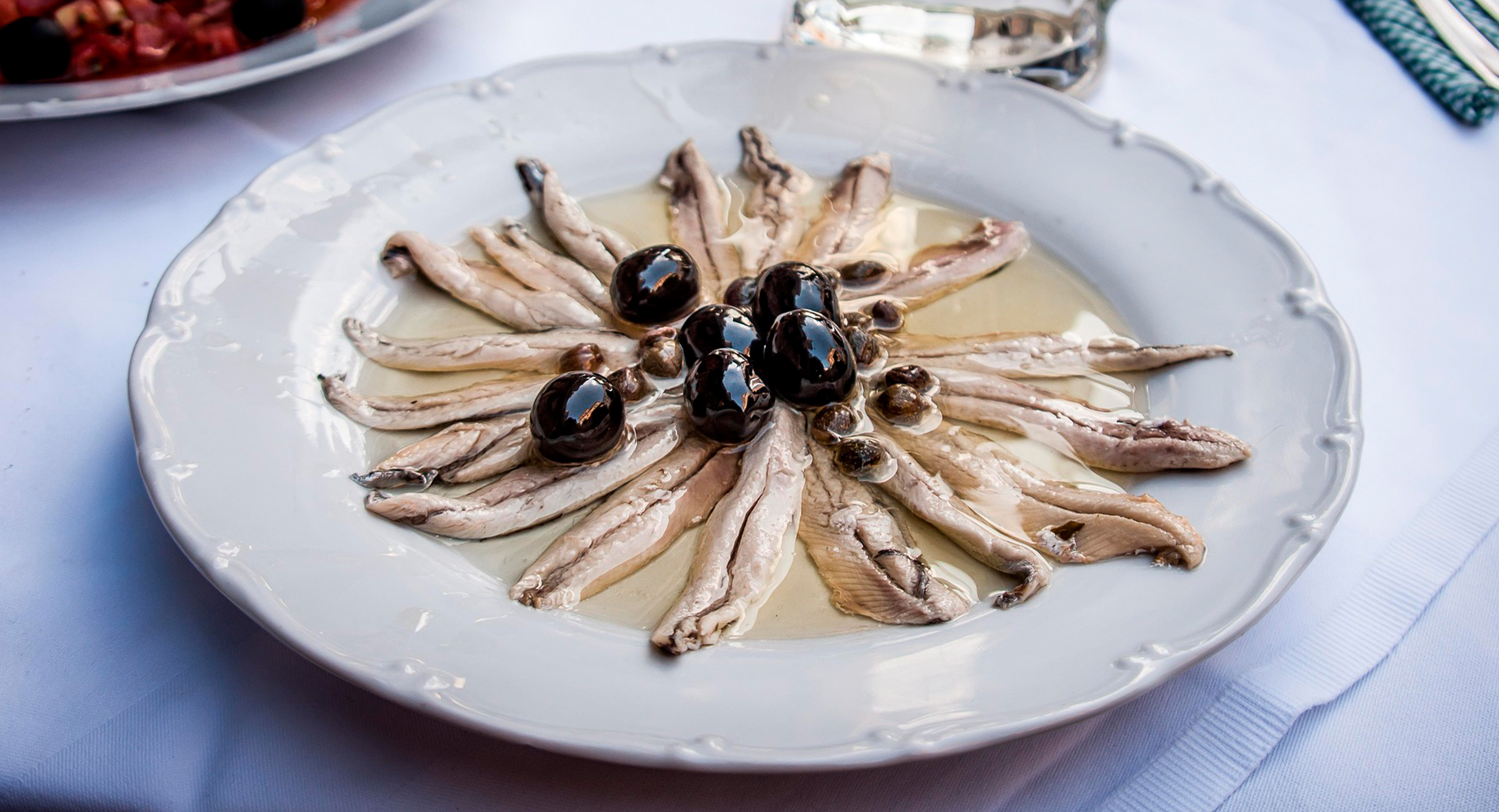 A imagem mostra um prato de anchovas servidas de forma elegante. Os filés estão dispostos em formato circular, como uma flor, sobre um prato branco, regados com bastante azeite. No centro, há azeitonas pretas e alcaparras, que complementam a apresentação.