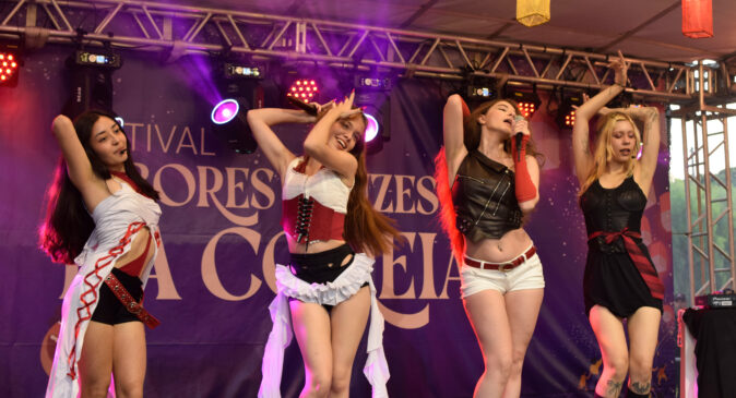 A imagem mostra quatro mulheres se apresentando em um palco, provavelmente durante um festival cultural. Elas estão dançando e cantando, com expressões animadas e poses coreografadas, enquanto luzes coloridas iluminam o cenário.