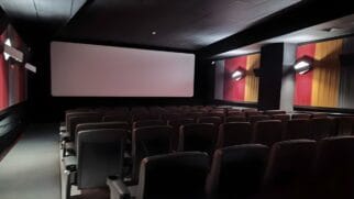 A imagem apresenta o interior de uma sala de cinema vazia, capturada de uma perspectiva lateral que mostra as fileiras de poltronas voltadas para a tela de projeção. O ambiente é predominantemente escuro, com paredes pretas que ajudam a concentrar a atenção na área de exibição. No centro da composição, destaca-se uma tela branca retangular e limpa, sem projeção no momento. As poltronas são de cor escura, possivelmente cinza ou pretas, organizadas em colunas e filas simétricas, equipadas com braços e suportes para copos. As paredes laterais possuem painéis decorativos verticais em tons de vermelho e amarelo, que quebram a monotonia do preto. Sobre esses painéis, há luminárias de parede em formato geométrico que emitem uma luz branca suave, criando pontos de brilho no ambiente. À esquerda, nota-se um corredor de circulação com uma pequena linha de luzes LED vermelhas no chão, indicando o caminho de segurança. O teto é escuro e liso, com pequenas saídas de ar e luzes embutidas discretas. A iluminação geral é baixa, típica de uma sala de cinema preparada para o início de uma sessão ou após o encerramento das atividades.