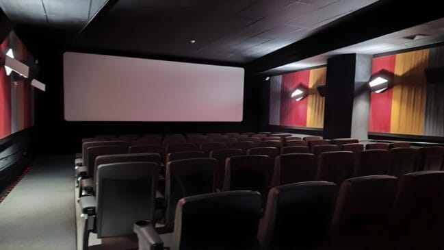 A imagem apresenta o interior de uma sala de cinema vazia, capturada de uma perspectiva lateral que mostra as fileiras de poltronas voltadas para a tela de projeção. O ambiente é predominantemente escuro, com paredes pretas que ajudam a concentrar a atenção na área de exibição. No centro da composição, destaca-se uma tela branca retangular e limpa, sem projeção no momento. As poltronas são de cor escura, possivelmente cinza ou pretas, organizadas em colunas e filas simétricas, equipadas com braços e suportes para copos. As paredes laterais possuem painéis decorativos verticais em tons de vermelho e amarelo, que quebram a monotonia do preto. Sobre esses painéis, há luminárias de parede em formato geométrico que emitem uma luz branca suave, criando pontos de brilho no ambiente. À esquerda, nota-se um corredor de circulação com uma pequena linha de luzes LED vermelhas no chão, indicando o caminho de segurança. O teto é escuro e liso, com pequenas saídas de ar e luzes embutidas discretas. A iluminação geral é baixa, típica de uma sala de cinema preparada para o início de uma sessão ou após o encerramento das atividades.