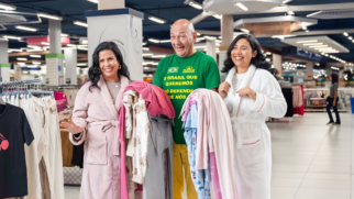 A imagem captura um momento descontraído e bem-humorado no interior de uma loja de departamentos, onde o empresário Luciano Hang aparece ao centro, vestindo sua tradicional camiseta verde com frases patrióticas, ladeado por duas mulheres que sorriem abertamente para a câmera. À esquerda, a dubladora Guilene Conte segura uma pilha de roupas coloridas enquanto veste um roupão rosa, enquanto à direita, uma jovem também trajada com um roupão branco compartilha do clima festivo da cena. O cenário ao fundo revela a amplitude da loja, com araras de roupas, iluminação industrial e prateleiras organizadas, evidenciando o contexto de uma gravação publicitária voltada ao setor de varejo em um ambiente vibrante e acolhedor.