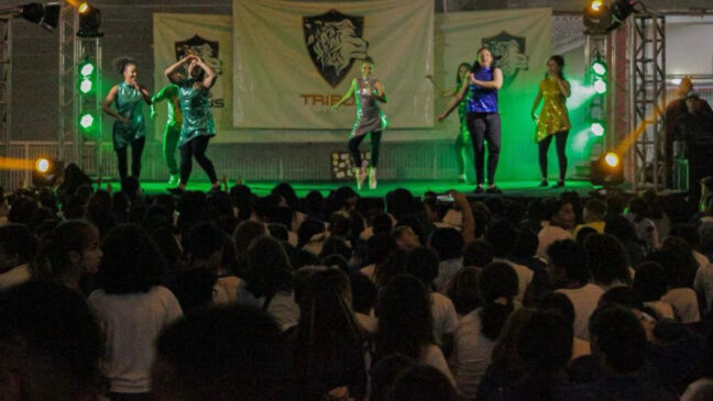 A imagem mostra uma apresentação de dança. Também é possível ver a plateia acompanhando o número de arte.