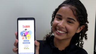 A imagem mostra uma jovem sorridente segurando um celular em primeiro plano. Na tela do aparelho, está aberto o aplicativo do programa ID Jovem, exibindo o logotipo oficial e as opções de acesso para o usuário. Ela tem cabelos cacheados e a foto possui um fundo neutro e claro