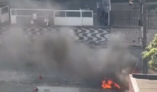 A imagem mostra um carro em chamas na rua, com fogo intenso concentrado na parte frontal do veículo. Uma grande quantidade de fumaça preta sobe e se espalha pelo local, reduzindo a visibilidade. Ao fundo, é possível ver algumas pessoas observando a cena à distância, próximas a uma calçada com padrão ondulado em preto e branco. A área parece urbana e movimentada, com muros, portões e estruturas ao redor. Não há presença visível de equipes de emergência no momento registrado da imagem.