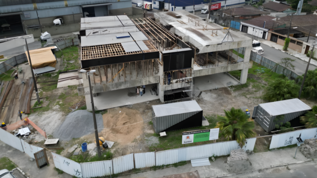 A fotografia aérea revela o estágio atual da construção da UME Hilda Rabaça, onde o esqueleto de concreto armado de dois pavimentos domina o centro do pátio em meio ao cenário urbano da Zona Noroeste. O edifício já apresenta lajes sólidas em algumas seções, enquanto outras partes ainda dependem de complexas estruturas de escoramento de madeira e vigas para as próximas etapas de concretagem. No pátio de terra batida que circunda a edificação principal, a logística da obra se manifesta através de contêineres metálicos que servem de suporte administrativo, pilhas de insumos como areia e brita, além de equipamentos essenciais como uma betoneira posicionada estrategicamente. O perímetro é rigorosamente isolado por tapumes metálicos brancos que se estendem ao longo da calçada, garantindo a segurança dos pedestres e a organização do espaço público. Próximo à entrada, a placa oficial da Prefeitura de Santos valida a transparência do projeto e identifica a intervenção para a comunidade local. Ao fundo, o entorno industrial e residencial, caracterizado por galpões e o tráfego de caminhões, reforça a inserção da nova unidade de ensino em uma área estratégica da Vila Haddad, evidenciando o contraste entre a robustez da nova estrutura e as edificações preexistentes do bairro