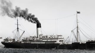 Imagem de época de um navio a vapor transatlântico em mar aberto. O navio tem múltiplos conveses, uma chaminé imponente com o topo preto e estruturas de mastros com cabos de aço. A foto tem granulação característica de registros do início do século XX, com o céu claro ao fundo.