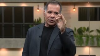 Uma fotografia em plano médio mostra o comediante e apresentador Leandro Hassum em um cenário de estúdio. Ele possui pele clara, cabelos grisalhos curtos e penteados para trás, vestindo uma camiseta preta sob um paletó cinza-escuro texturizado. O apresentador olha diretamente para a câmera enquanto faz um gesto com a mão direita, unindo as pontas do polegar e do indicador em um sinal de "ok" ou para enfatizar um ponto, mantendo os outros dedos levemente levantados. O fundo da imagem está suavemente desfocado, revelando um ambiente moderno com tons pastéis e amadeirados. É possível observar elementos decorativos como plantas verdes em jardineiras ao fundo e luminárias de filamento presas à parede, que emitem uma luz quente e suave. À esquerda, há uma estrutura que se assemelha a uma janela com molduras pretas. A iluminação frontal é clara e uniforme, destacando as expressões faciais do apresentador. A composição sugere um momento de fala durante uma transmissão de televisão ou evento.