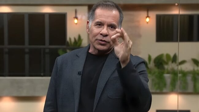 Uma fotografia em plano médio mostra o comediante e apresentador Leandro Hassum em um cenário de estúdio. Ele possui pele clara, cabelos grisalhos curtos e penteados para trás, vestindo uma camiseta preta sob um paletó cinza-escuro texturizado. O apresentador olha diretamente para a câmera enquanto faz um gesto com a mão direita, unindo as pontas do polegar e do indicador em um sinal de "ok" ou para enfatizar um ponto, mantendo os outros dedos levemente levantados. O fundo da imagem está suavemente desfocado, revelando um ambiente moderno com tons pastéis e amadeirados. É possível observar elementos decorativos como plantas verdes em jardineiras ao fundo e luminárias de filamento presas à parede, que emitem uma luz quente e suave. À esquerda, há uma estrutura que se assemelha a uma janela com molduras pretas. A iluminação frontal é clara e uniforme, destacando as expressões faciais do apresentador. A composição sugere um momento de fala durante uma transmissão de televisão ou evento.