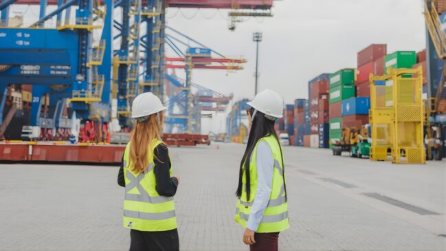 A imagem mostra duas mulheres de costas, vestindo capacetes brancos e coletes refletivos amarelos, observando a operação de um terminal portuário. Elas estão em pé sobre um piso de paralelepípedos cinza claro que se estende ao fundo. À esquerda, há grandes guindastes portuários (portêineres) em azul e amarelo, utilizados para a movimentação de cargas. À direita, fileiras de contêineres coloridos (vermelho, verde, azul e laranja) estão empilhados de forma organizada. O cenário é de um ambiente industrial e logístico dinâmico, sob um céu nublado e claro.