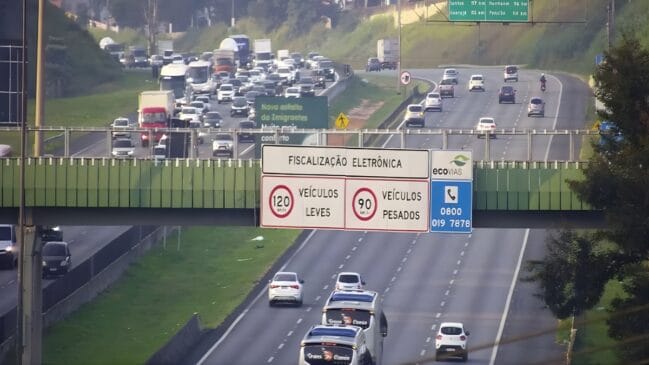 A imagem registra o fluxo de veículos em um trecho de rodovia do Sistema Anchieta-Imigrantes durante o dia. Em primeiro plano, uma passarela verde atravessa a estrada, sustentando uma grande placa branca de fiscalização eletrônica que indica os limites de velocidade: 120 km/h para veículos leves e 90 km/h para veículos pesados. Ao lado direito da placa, há um painel azul com o logotipo da Ecovias e o número de emergência 0800. Sob a passarela, carros e ônibus brancos circulam por três faixas de asfalto em direção ao observador. Ao fundo, no sentido oposto da via, observa-se um congestionamento intenso com diversos automóveis, caminhões e carretas parados ou em movimento lento, ladeados por encostas gramadas. No canto superior direito, uma placa de sinalização aérea verde indica distâncias para cidades como Santos, Guarujá, Itanhaém e Peruíbe. A iluminação é natural, sugerindo uma manhã ensolarada com uma leve névoa ao horizonte.