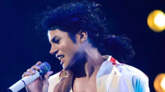 A imagem mostra um cover de Michael Jackson caracterizado com o visual icônico do cantor, segurando um microfone e cantando em um palco iluminado. Ele usa o clássico cabelo cacheado e uma camisa branca aberta.