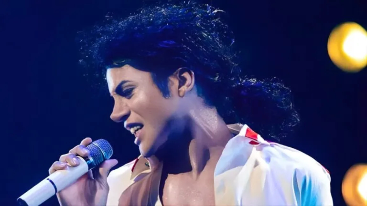 A imagem mostra um cover de Michael Jackson caracterizado com o visual icônico do cantor, segurando um microfone e cantando em um palco iluminado. Ele usa o clássico cabelo cacheado e uma camisa branca aberta.