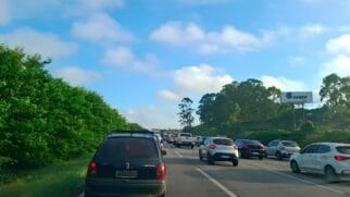 A imagem retrata um congestionamento em uma rodovia sob um céu azul com nuvens esparsas. Em primeiro plano, à esquerda, destaca-se a traseira de um Chevrolet Corsa preto. À direita, diversos veículos, incluindo modelos como o Renault Kwid e o Sandero, ocupam as faixas de rolagem em fluxo lento. As margens da estrada são compostas por uma vegetação densa e verde, e ao fundo, no lado direito, observa-se um grande outdoor da empresa Sanko instalado em uma área elevada e arborizada.