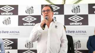 Homem de camisa branca falando ao microfone em evento com painéis do Santos FC e da Umbro ao fundo.
