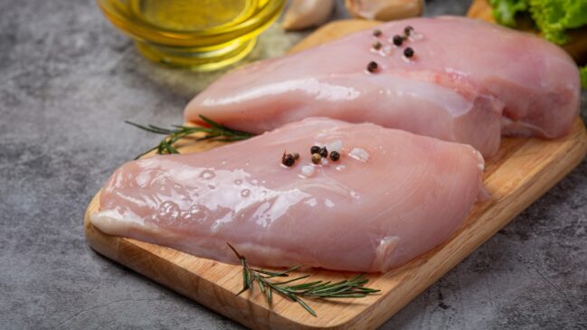 Dois filés de peito de frango crus temperados com pimenta em grão sobre tábua de madeira.