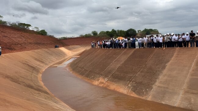 Comitiva de autoridades e trabalhadores reunida na margem de um canal artificial de água sob céu nublado.
