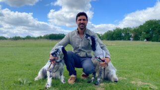 Homem agachado em um gramado verde ao ar livre, segurando dois cães da raça Setter Inglês pelas coleiras. Ele veste uma jaqueta cinza e calças escuras, sorrindo para a câmera. Ao fundo, há uma vasta área de campo com árvores e um céu azul ensolarado com nuvens brancas