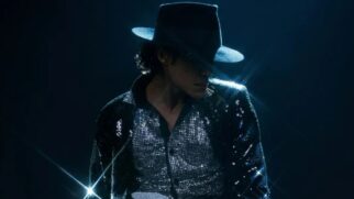 Jafaar Jackson interpretando Michael Jackson inclinado com chapéu cobrindo os olhos e jaqueta de lantejoulas refletindo luzes intensas.